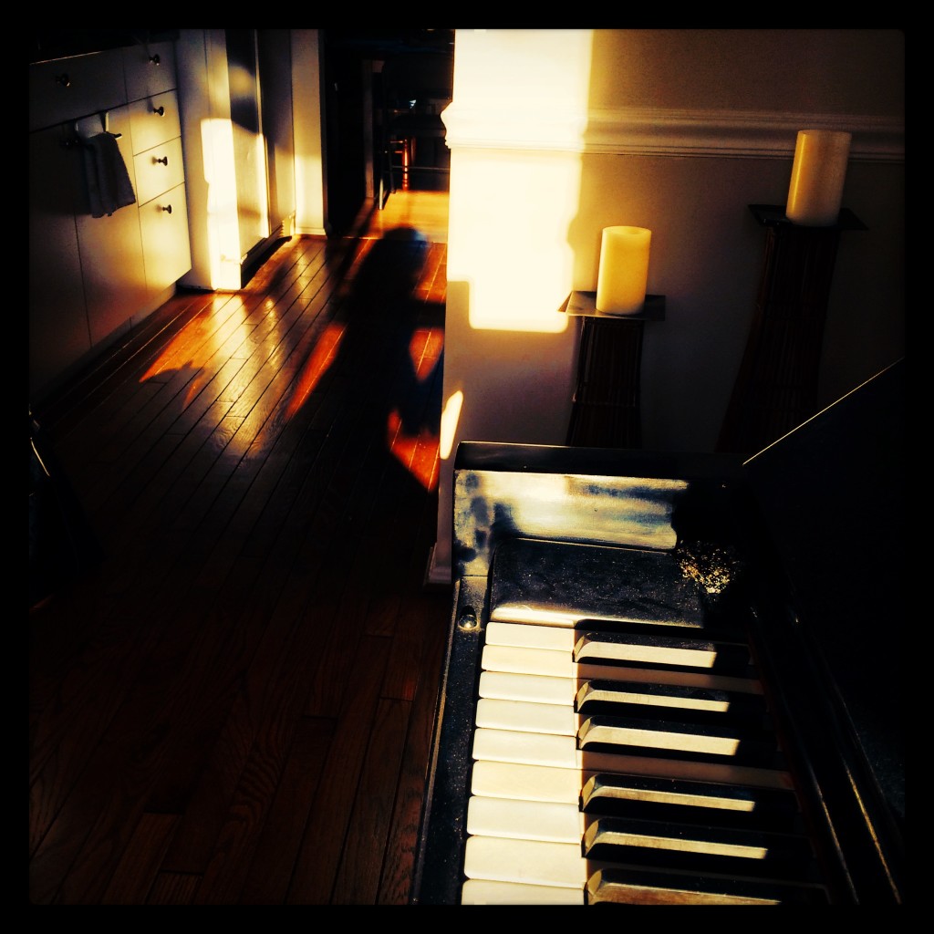 Image description: A “selfie” (self portrait) of my shadow silhouette sitting down during an improv session with my Steinway upright piano in the Front room of my home, looking out through the kitchen and dining area. The photo was captured at sunset.