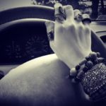 Image description: a white cisgender female hand placed on a steering wheel inside a parked car. The hand is decorated with rings and bracelets.