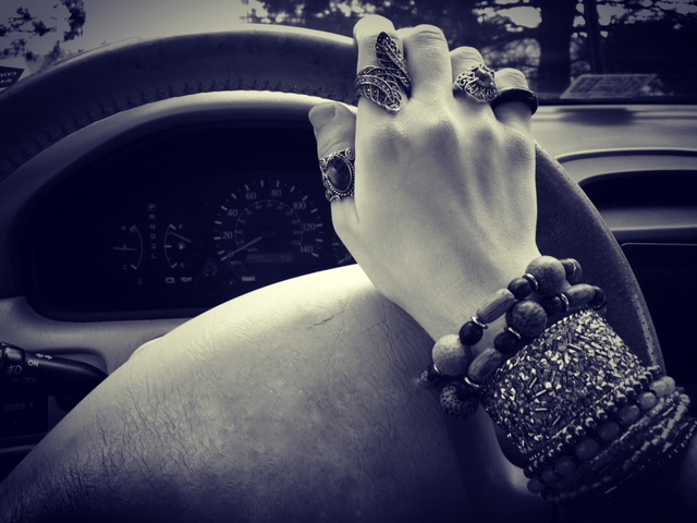 Image description: a white cisgender female hand placed on a steering wheel inside a parked car. The hand is decorated with rings and bracelets.
